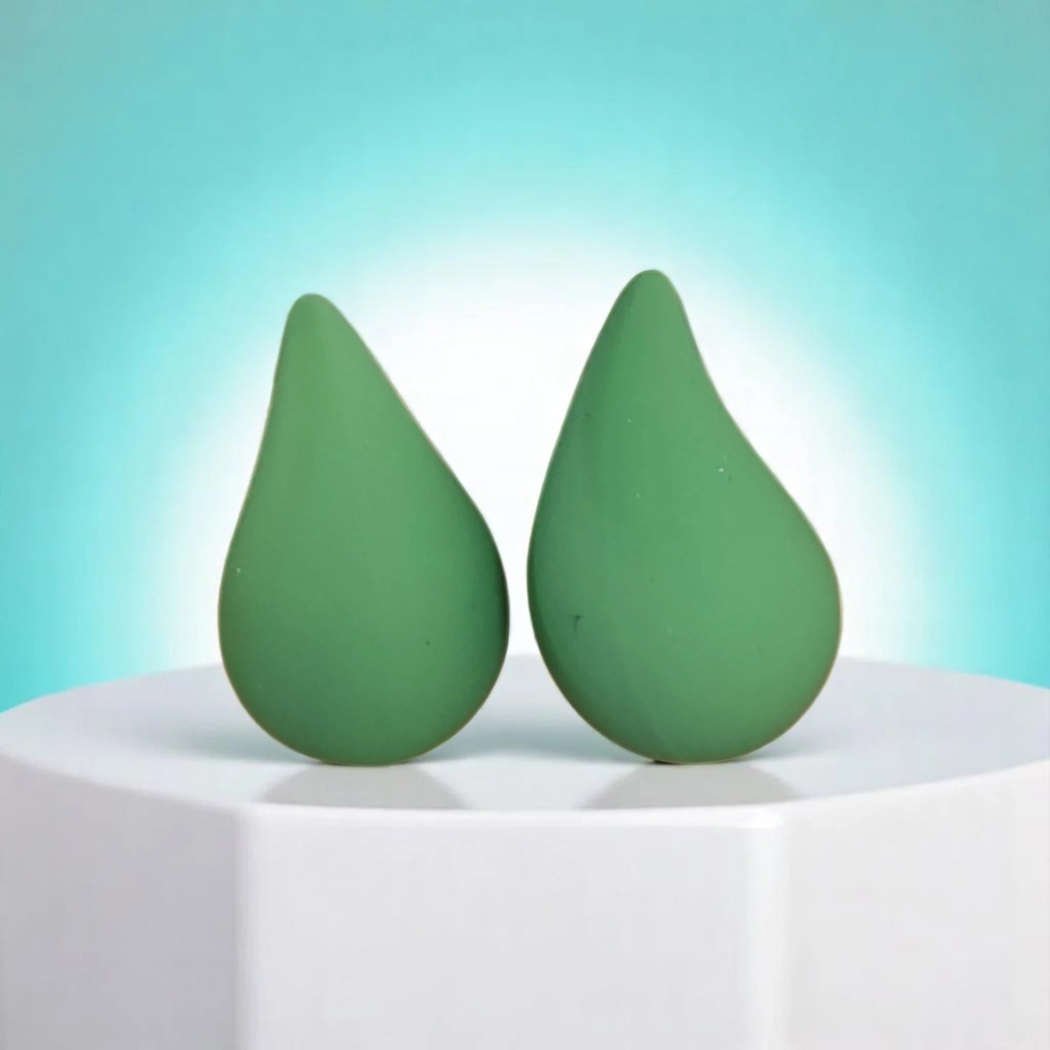Two green, teardrop-shaped beauty blenders displayed on a white pedestal against a light blue background.