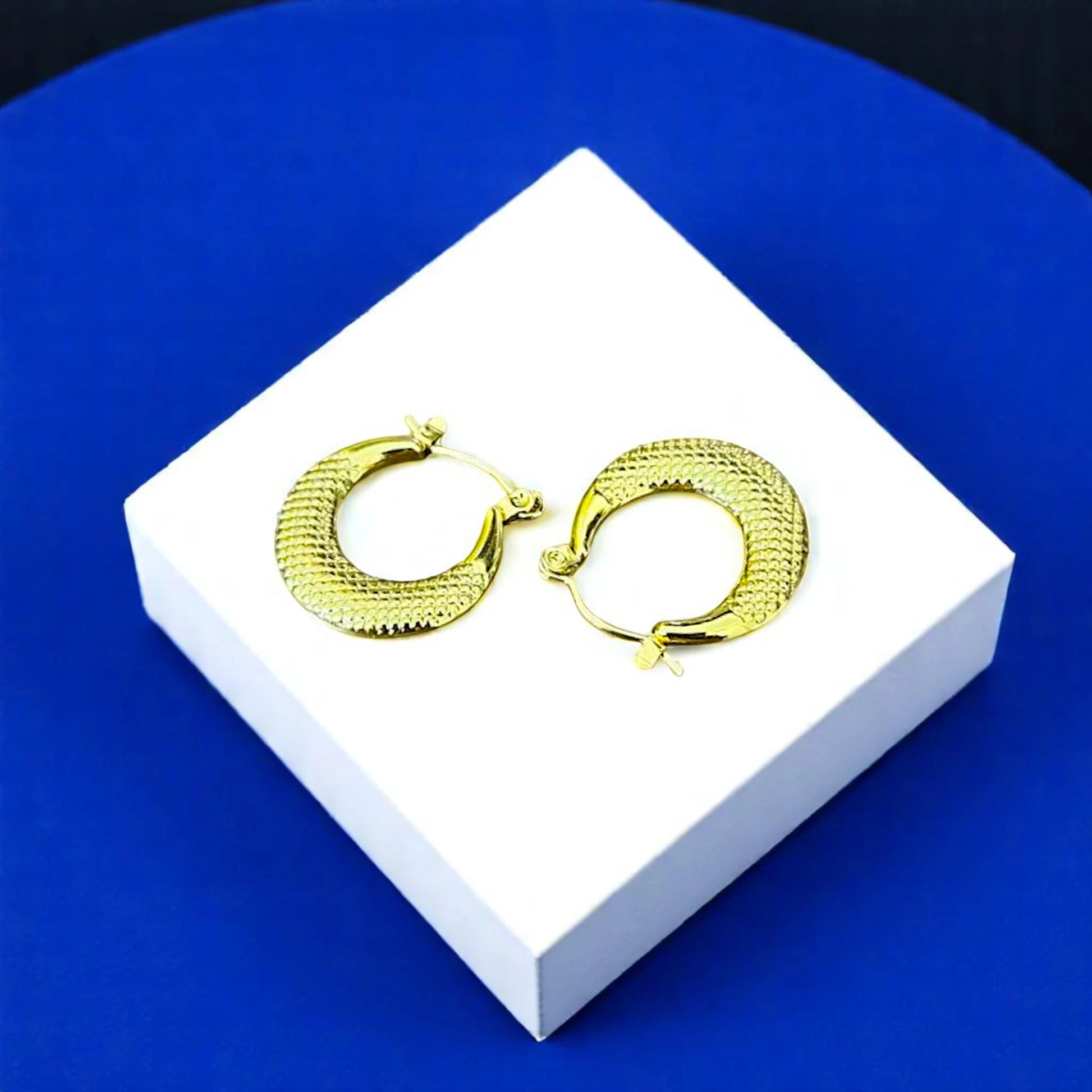 Gold hoop earrings with a textured design displayed on a white box against a blue background.