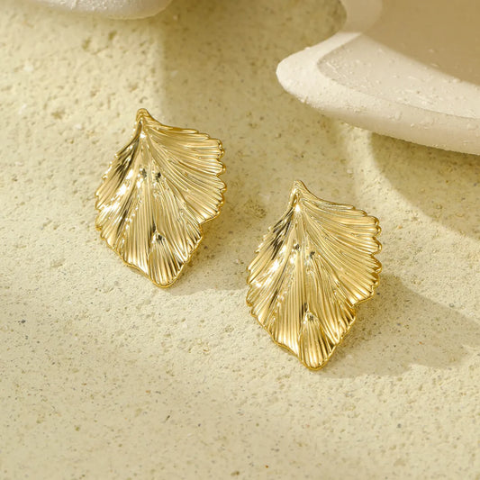 Delicate gold leaf-shaped earrings displayed on a textured sandy surface, showcasing intricate detailing and a lustrous finish.