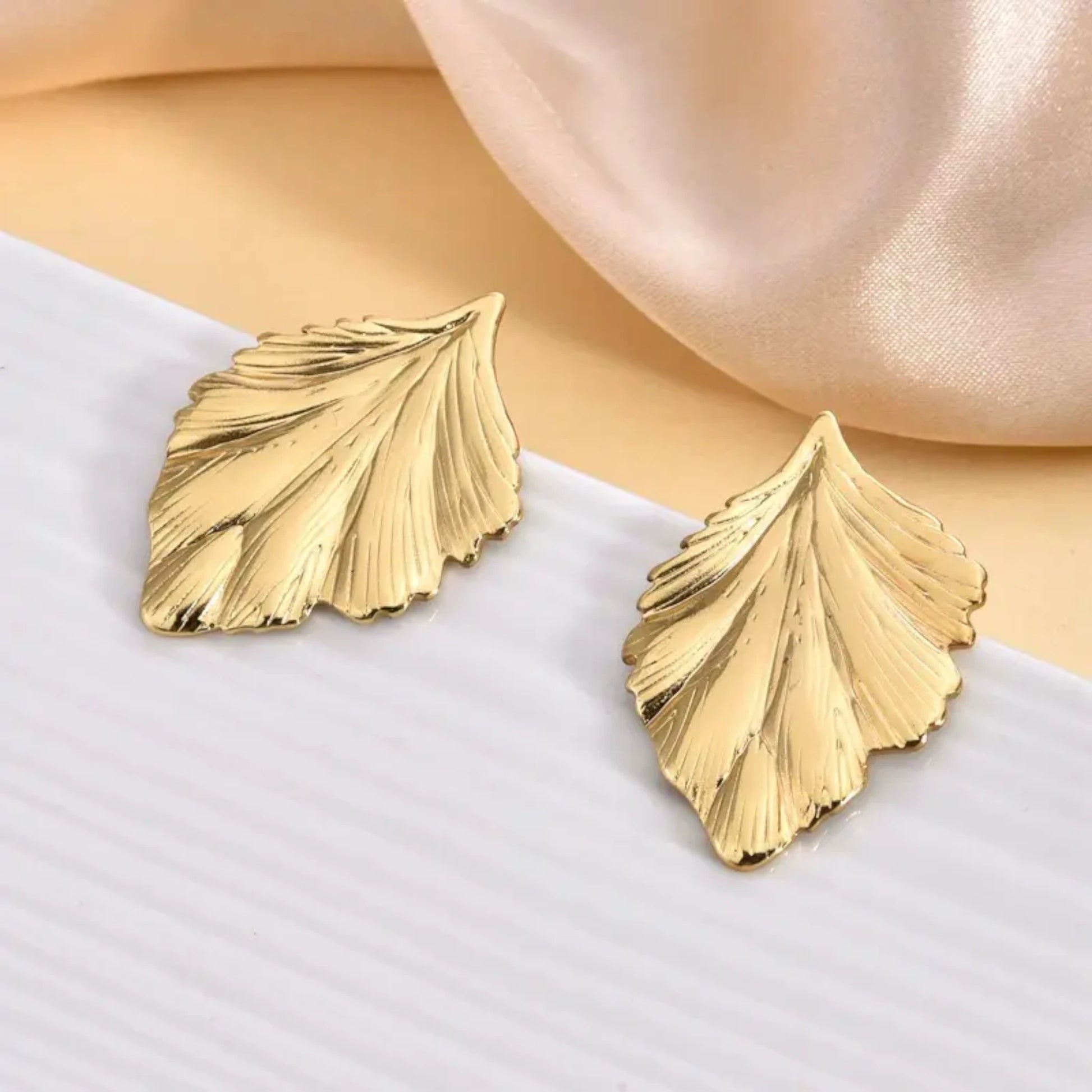 Gold leaf-shaped earrings displayed on a textured surface, with soft fabric in the background, showcasing their elegant design.