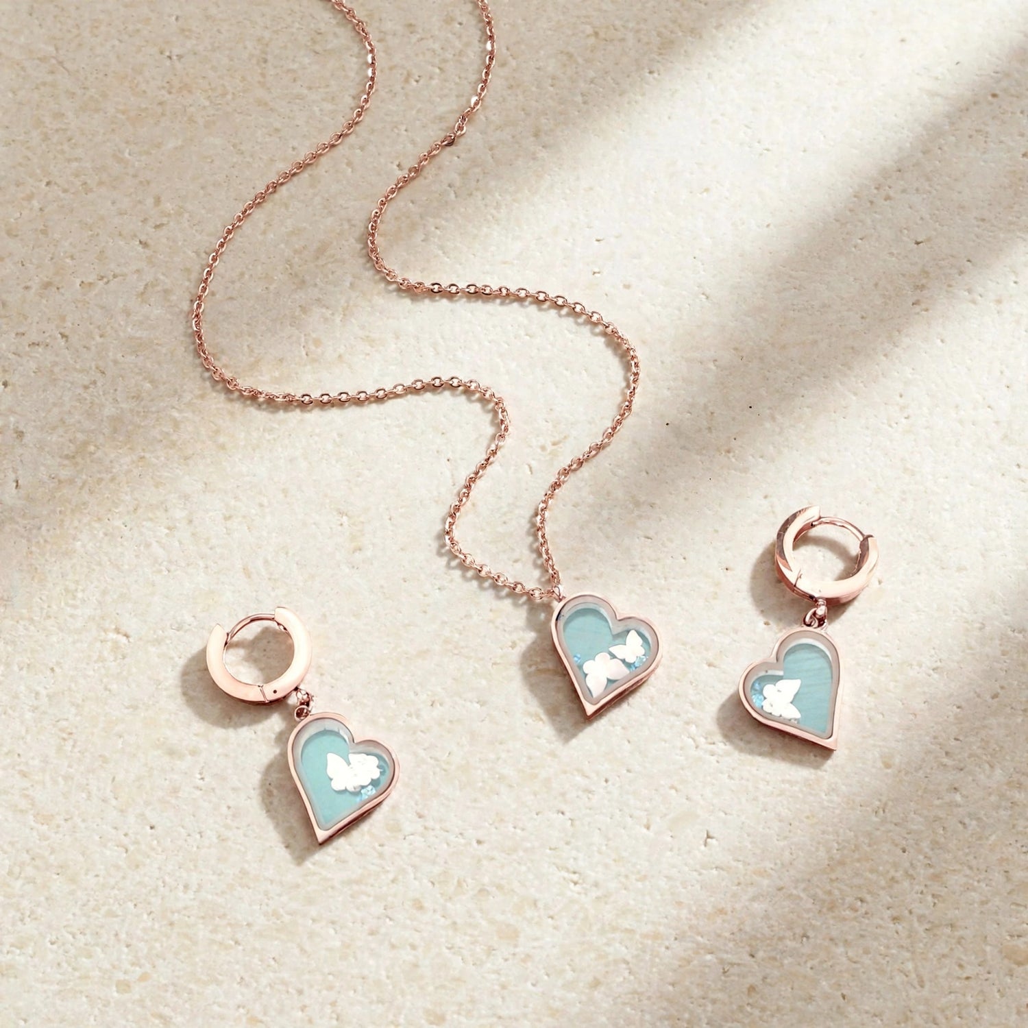 Rose gold jewelry set featuring a delicate chain necklace, a matching drop earring, and a small hoop earring, each with a heart-shaped charm filled with a light blue and white floral pattern, displayed on a textured beige surface with soft shadows.