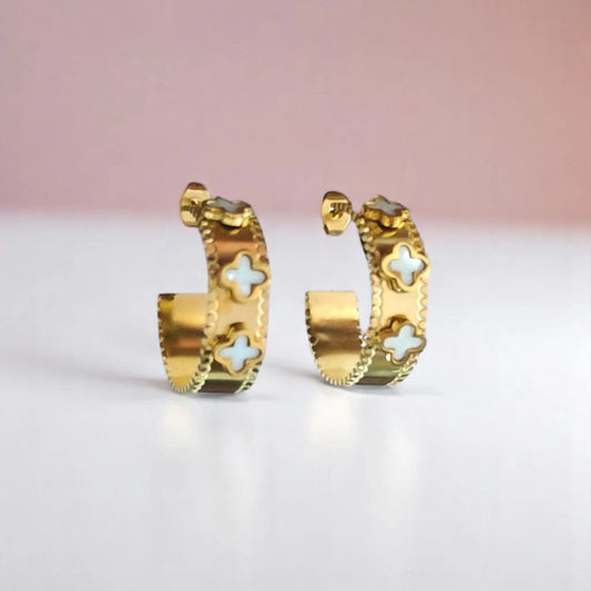 Gold hoop earrings featuring intricate designs and small light blue cross embellishments, set against a soft pink background.