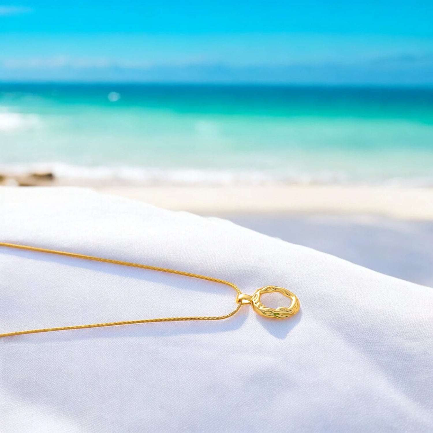 Elegant gold wavy circle pendant necklace resting on white fabric with a serene beach and ocean backdrop. Perfect for minimalistic and beach-inspired jewelry lovers.