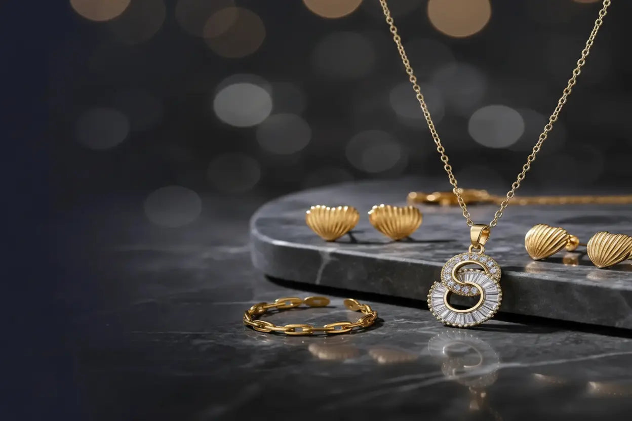 Gold jewelry set featuring a pendant necklace with a stylized letter 'S' pendant, a matching ring, and shell-shaped earrings, displayed on a dark marble slab with a dark blurred background.