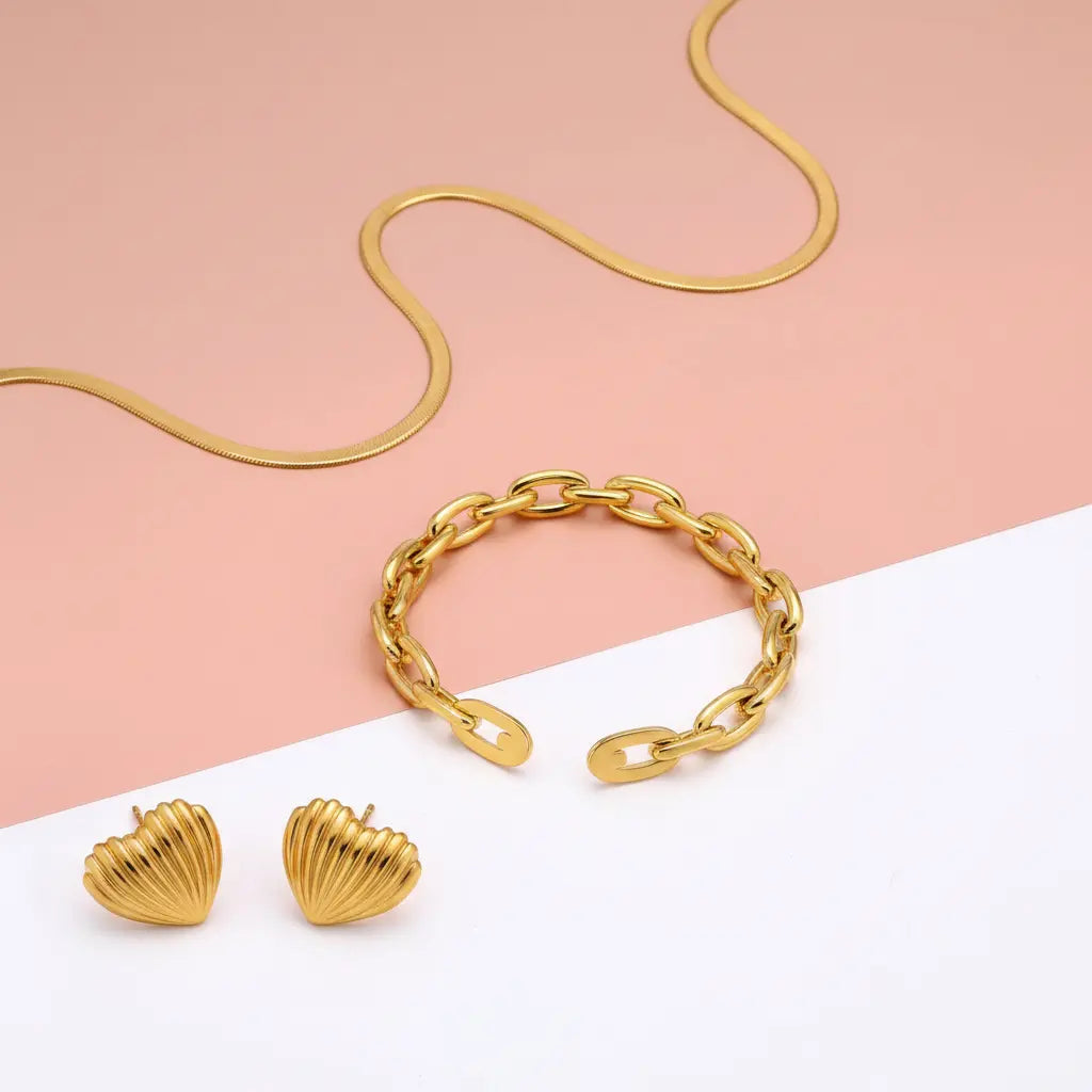 Gold jewelry set featuring a chain bracelet, matching chain necklace, and two ribbed heart stud earrings, arranged on a split pink and white background.