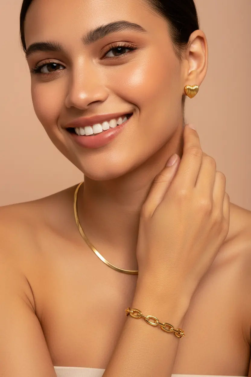A smiling woman wearing a gold chain necklace, a gold link bracelet, and a small gold heart-shaped stud earring, posed against a warm beige background.